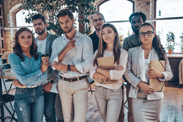 Group of young modern people in smart casual wear looking at camera while standing in the office