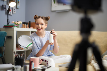 Smiling teen girl holding bottle of cream and recording video blog at home