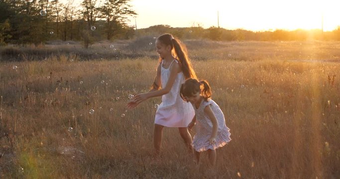 Two little pretty sisters, four and seven years old, have fun catching big soapy air bubbles in a  meadow near the forest on a  summer sunset. Slow motion 4k 50 FPS