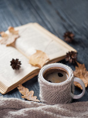 Fototapeta na wymiar A mug of coffee in a knitted vest or a handmade sweater and an old book on the table. Autumn composition with a mug of hot coffee and autumn dry leaves. Home comfort. Hygge concept