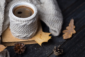 A mug of coffee in a knitted vest or a handmade sweater and an old book on the table. Autumn composition with a mug of hot coffee and autumn dry leaves. Home comfort. Hygge concept