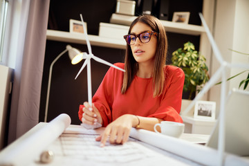 Dedicated hardworking pretty businesswoman dressed in formal wear sitting in office and holding windmill model. On table are blueprints and laptop. Sustainable development concept.