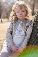 pretty curly kid girl in leather jacket posing near big tree in the city park alone