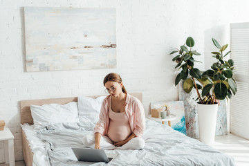 pregnant woman in glasses sitting on bed and using laptop