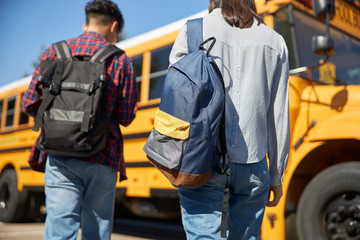 Two teenagers are holding backpack near school bus