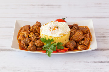 Pork chop with tomato sauce, polenta and fried egg on white plate on white wood background