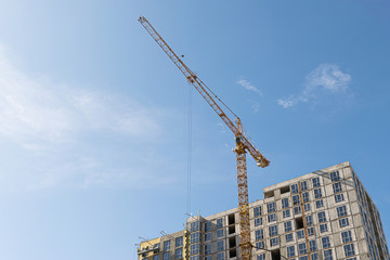 A yellow high-rise building crane against a blue sky builds multi-storey apartment buildings using modern technologies of metal, concrete and brick according to an architectural project
