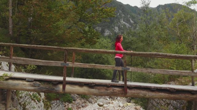 Drone ascending over a small wooden bridge, woman hiking, walking over from left to right, mountains and forest in background.