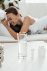 selective focus of suffering woman in bed and pills with glass of water on foreground