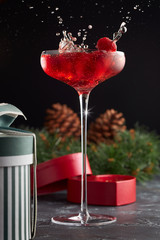 Glass of pink champagne on a high stem close - up with splashes on the background of boxes with gifts and a Christmas wreath with cones. Festive still life.