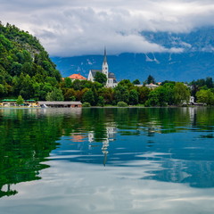 Fototapeta na wymiar View of Lake Bled and St. Martin's Church with reflection. Clouds cover the tops of the mountains.