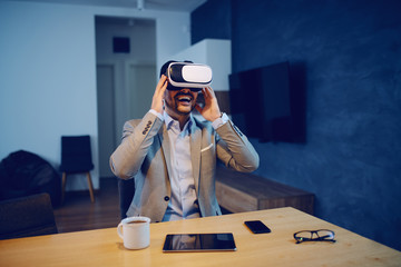 Handsome smiling Caucasian unshaven man in suit sitting in dining room in morning and using virtual reality headset. On table are eyeglasses, smart phone, tablet and coffee.
