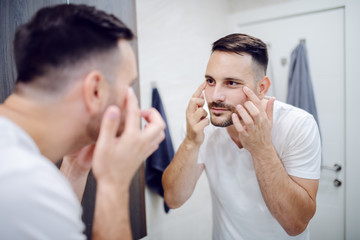 Mirror reflection of good looking Caucasian man checking his wrinkles under eyes. Male cosmetics concept.