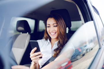 Gorgeous caucasian brunette smiling brunette sitting in her car with safety belt and using smart phone for texting message.