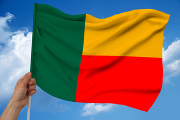 hand holds against the background of the sky with clouds the colored flag of Benin on the texture of the fabric, silk with waves, close-up