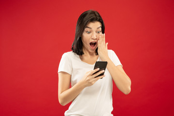 Naklejka premium Caucasian young woman's half-length portrait on red studio background. Beautiful female model in white shirt. Concept of human emotions, facial expression. Buys online, payment bill, looks wondered.