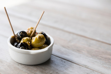 black and green olives in white bowl with wooden stick. On white table, space for text