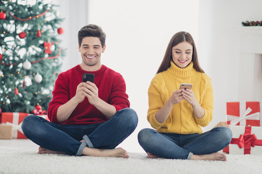 Full length photo of two cheerful romantic people sit on floor use cellphone type congratulations with newyear search christmas season discounts in house with x-mas decoration