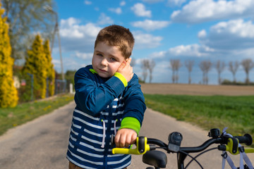 Five-year cute boy standing on the street and holding his ear, a small bicycle visible and the sky with clouds.