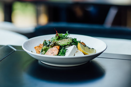 Grilled Salmon With Mixed Salad And Avocado On Table With Black Table Cloth In Fine Dining Restaurant With Window Light.