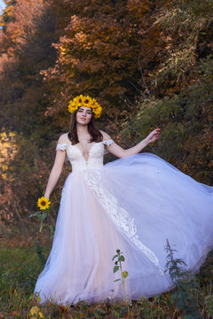 Beautiful cute sexy girl in a white dress with a wreath of sunflowers on her head enjoying nature. Autumn time.