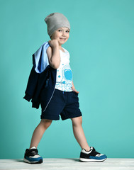 Cool naughty kid boy in blue shorts, white shirt and hat is walks carrying his hoody in his hand over his shoulder