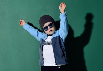 Portrait of cool kid boy in blue sunglasses, hat, fleece jacket and white t-shirt having fun jumping dancing on green