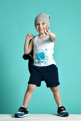 Happy smiling kid boy in blue shorts, white shirt, hat and jacket he is holding on his shoulder is showing V sign