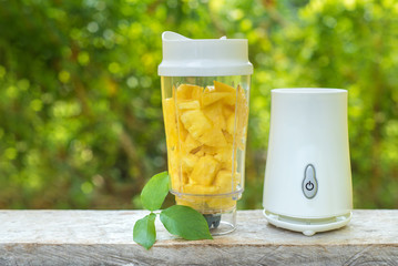 A white blender with pineapple slices inside stands on a wooden table against the summer foliage....