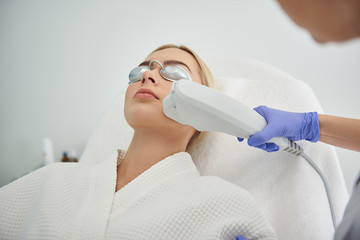 Young lady in goggles receiving laser facial treatment in beauty salon