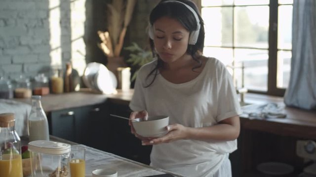 Medium shot of young mixed-race woman standing at kitchen table, eating cereals and listening to music through headphones and then starting singing using spoon as microphone