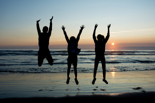 Silhouettes of three teanagers jumping out joyfully on the beach with setting sun at the bakcground