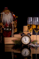 Two wine glasses with champagne, Santa Claus, clock and christmas gifts on a black background with reflection