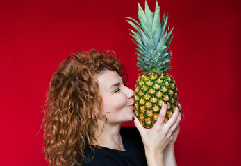 Red-haired beaut in a black t-shirt with a print of heart hold a pineapple in her hands on a red background. Happy girl. Amazing emotions. Sweet pineapple.