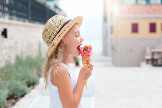 Fototapeta Kid is eating ice cream. Little girl is enjoying melting italian gelato. Happy cute child in straw hat is tasting delicious food in town streets. Concept of childhood, lifestyle moments, summer travel