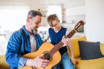 Mature father with small son sitting on sofa indoors, playing guitar.