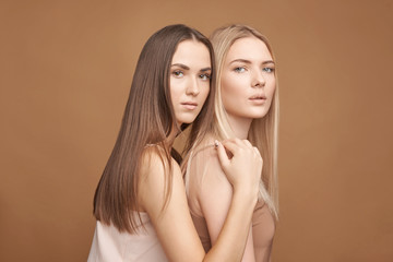 Two beautiful young women with a different skin type dressed in tops of natural color on a brown background, natural makeup, beautiful radiant skin, long shiny hair.