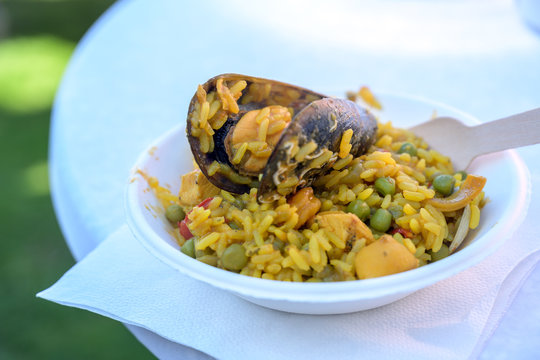 Fototapeta spanish street food, paella of rice with seafood and mussel in a sustainable cardboard bowl and a wooden fork on a white table, close-up shot