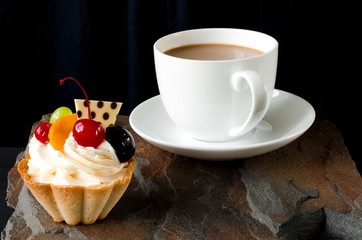 Fruit cake with cherries, grapes and plum stands on a wild stone next to a white cup of coffee with a foam