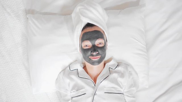 female with a white towel on her head and black cosmetic mask lying on the bed holding painted smiley have fun at home wellness, spa, skin care