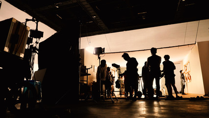 Behind the shooting video production and lighting set for filming which movie crew team working and silhouette shadow of camera and professional equipment in big studio for commercial advertising. - obrazy, fototapety, plakaty