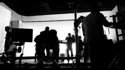 Behind the shooting video production and lighting set for filming which movie crew team working and silhouette shadow of camera and professional equipment in big studio for commercial advertising.