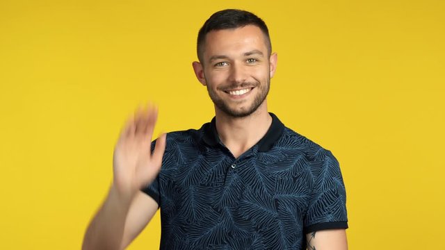 Handsome happy man waving hello or goodbye to camera on yellow background