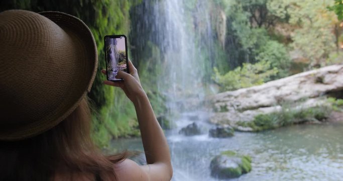 Rear view of woman in hat taking photograph smartphone sharing photo of waterfall nature background.