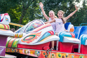 Happy mother and daughter, like sisters, riding the merry-go-round together. With the speed of going on a roller coaster, laughing, smiling, waving cheerfully while spending time in an amusement Park.