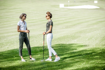 Two female best friends walking with golf putters on beautiful playing course, talking and having fun during a game on a sunny day