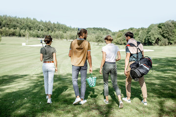 Young and elegant friends walking with golf equipment, hanging out together before the golf play on the beautiful course on a sunny day, rear view