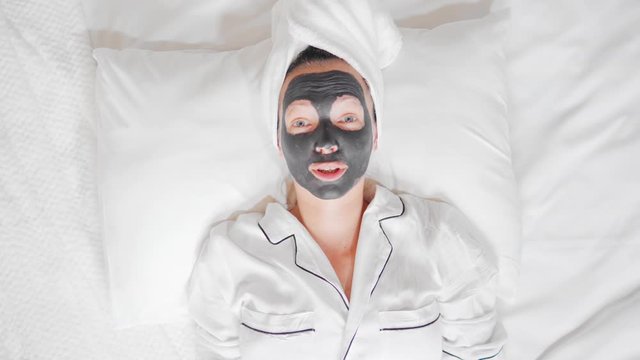 woman with a white towel on her head and black cosmetic mask lying on the bed using webcam for blogging talking with viewers at home wellness, spa, skin care
