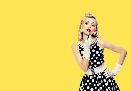 Young woman in pin up style black and white dress in polka dot, applying lipstick, isolated over yellow color background