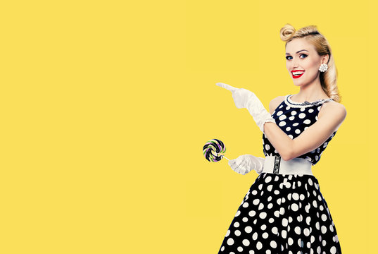 Smiling blond woman in pin up style in black and white dress in polka dot, showing something or empty copy space area for some text, slogan or advertising, isolated over yellow color